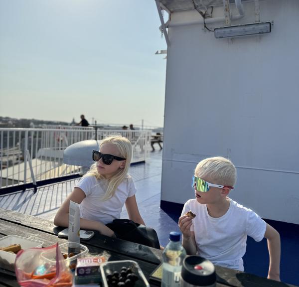 Två blonda barn med solglasögon sitter på ett soldäck på en färja 